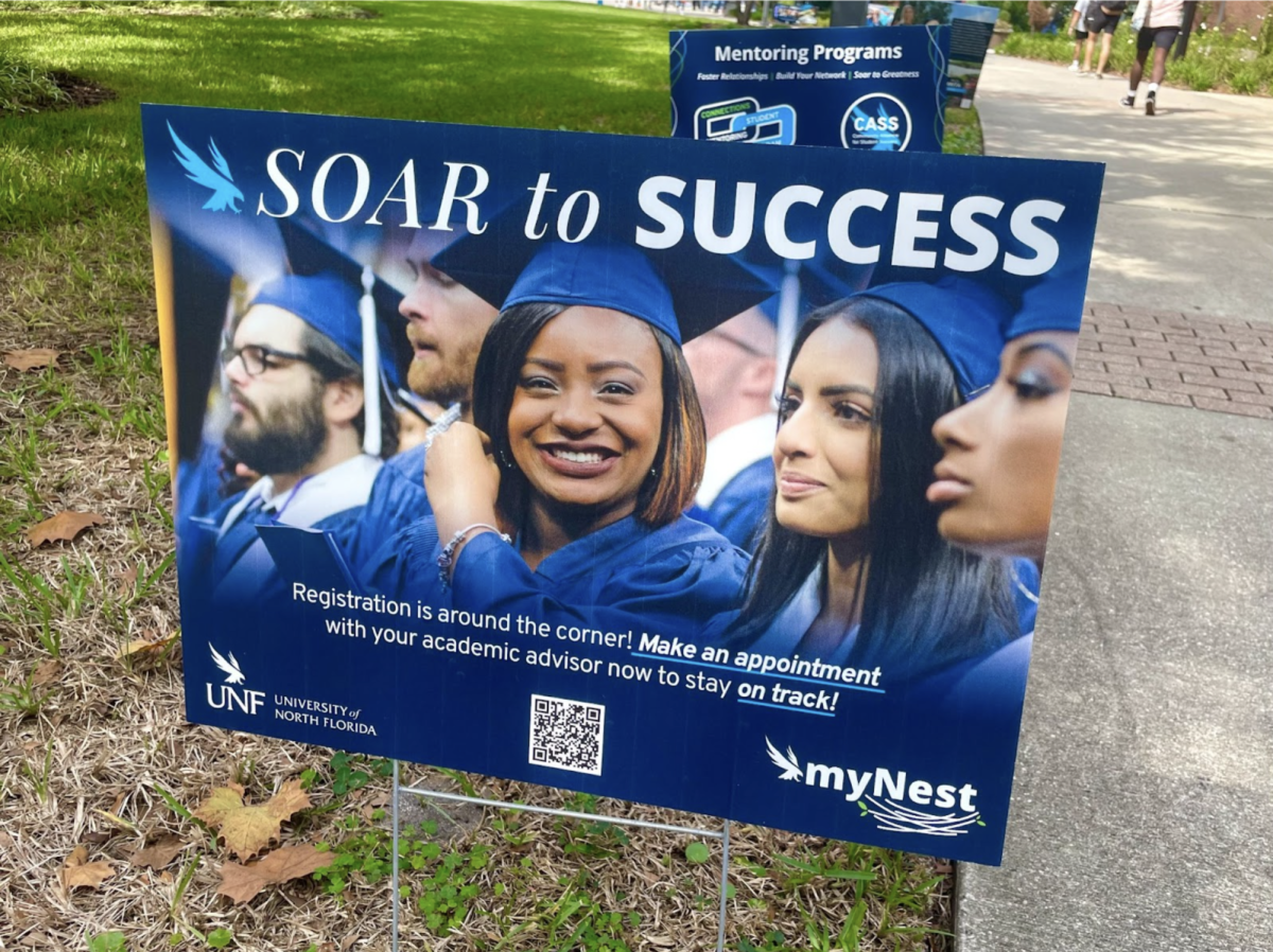 A blue UNF yard sign with the words "Soar to Success" and a photo of graduating students sits in the grass next to a sidewalk, encouraging students to register for fall and summer classes.